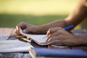 A person sits at an outdoor table, holding a pen and open book, with another book—A Buyer's Guide to Distance Learning—lying nearby. Sunlight creates a warm, natural setting as the person’s hands focus on studying or taking notes.