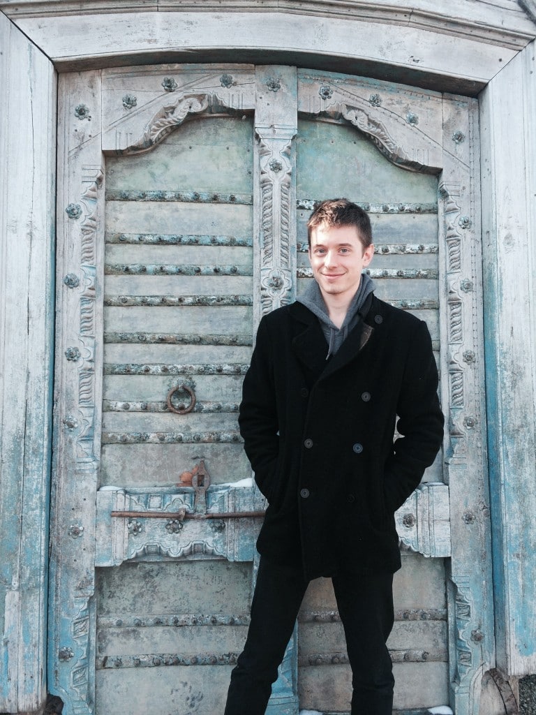 Brandon George, in a black coat, stands smiling in front of an ornate, weathered wooden door with intricate carvings and metal details.