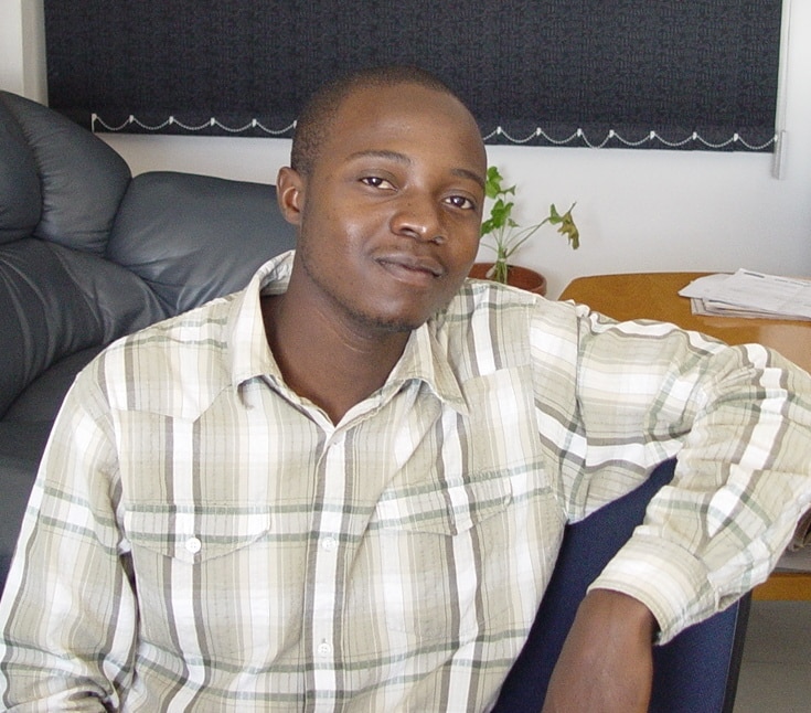 A man, Brian Madzokere, wearing a light checked shirt sits indoors, resting his arm on a chair. Behind him are a grey sofa, a small wooden table with papers, and a potted plant. He looks at the camera with a relaxed expression.