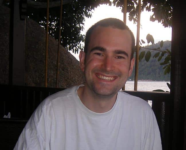 Chris Barraclough, a man with short dark hair and a white T-shirt, smiles at the camera whilst sitting outdoors near a wooden railing, with trees and water visible in the background.