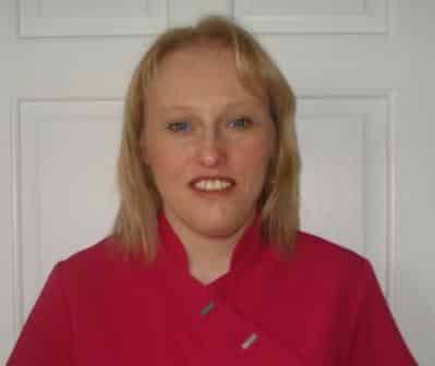 Clare Pearshouse, a woman with blonde hair wearing a bright pink top, stands smiling in front of a white panelled door.