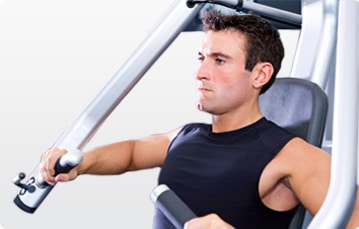 A man wearing a sleeveless black shirt is using a chest press machine at the gym, gripping the handles and pushing forward with a focused expression.