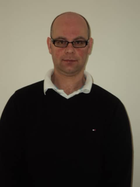 Thomas Ian Surgenor, a man with glasses wearing a dark jumper over a white collared shirt, stands in front of a plain light-coloured wall and looks at the camera with a neutral expression.