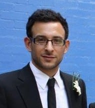 Ian Shine, wearing glasses and a black suit with a white shirt and black tie, stands in front of a blue brick wall. He has short dark hair and a buttonhole on his lapel.