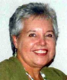 Janice Speddings, a woman with short grey hair, wearing a green top and earrings, smiles at the camera against a plain light background.