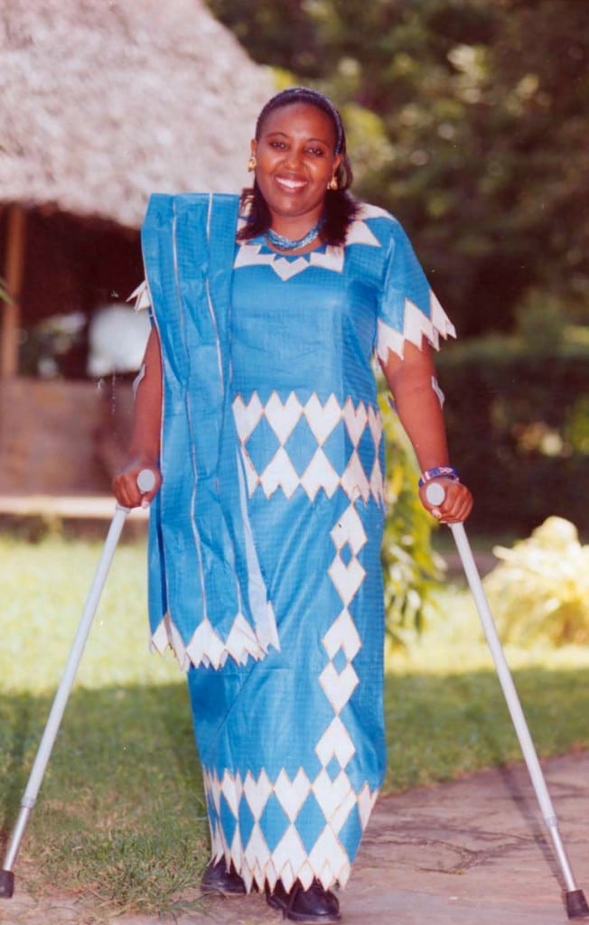 Jemimah Kutata, wearing a blue and white patterned dress, stands outdoors smiling at the camera and holding crutches. There are trees and a thatched roof in the background.