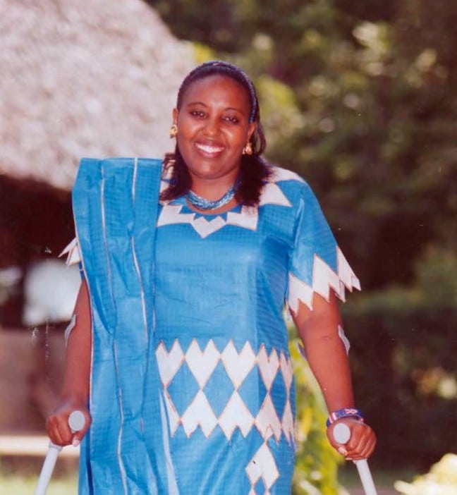 Jemimah Kutata, wearing a blue and white patterned dress, stands outdoors smiling at the camera and holding crutches. There are trees and a thatched roof in the background.