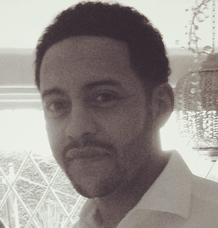 A man with short curly hair, a trimmed beard, and moustache, wearing a white collared shirt, stands indoors near a window with patterned glass. The photo is in black and white.