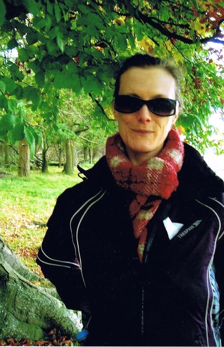 Katy Long, wearing dark sunglasses, a knitted scarf, and a dark jacket, stands outdoors under a leafy tree, with green grass and additional trees in the background.