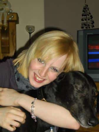Kayleigh Ashford, a smiling woman with blonde hair, hugs a large black dog. She is wearing a bracelet and a scarf. In the background, there is a television and a small decorative tree.
