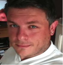 Lee Oakley, a man with short dark hair and green eyes, wearing a white shirt, is looking at the camera and smiling slightly. He has a stud earring in his left ear, and the background appears to be indoors.