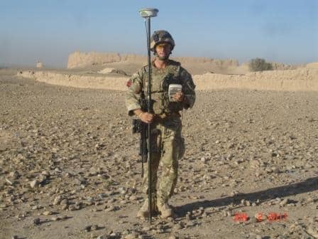 A soldier from the Martin Pyle Royal Commando Engineers, in camouflage uniform and helmet, stands in a rocky desert holding surveying equipment with a GPS receiver. Ancient ruins rise in the background beneath a clear sky.