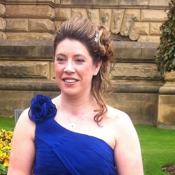 A woman with wavy brown hair styled up, wearing a one-shoulder royal blue dress with a large flower detail, smiles outdoors in front of a stone building and green lawn—capturing Nichola Learie’s radiant elegance.