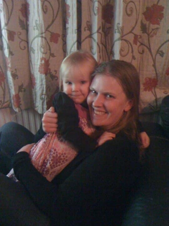 Nicola Halliday, a woman with long hair, smiles whilst hugging a young child in a pink dress, sitting together in front of floral-patterned curtains.