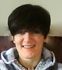 Nicola Rowan-Brooks, with short dark hair, is smiling whilst wearing a light-coloured hooded jumper and sitting against a plain background.