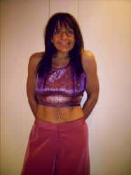 Ria Cohen, a woman with long dark hair, smiles at the camera. She is wearing a sleeveless, colourful crop top and loose, pink trousers, standing in front of a plain, light-coloured background.