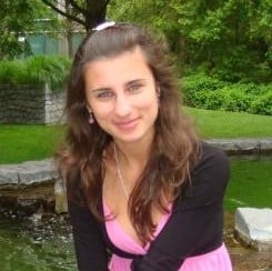 Rodica Petrea, a young woman with long brown hair, wearing a pink top and black cardigan, sits in front of a pond in a green park, smiling at the camera.
