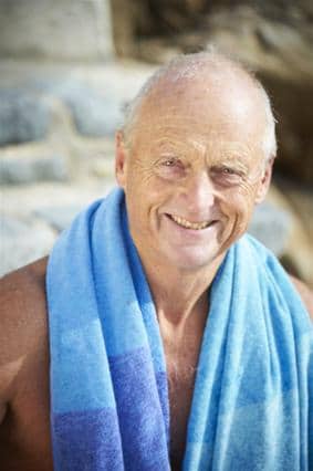 Roger Allsopp, an older man with short grey hair, smiles at the camera, wearing a blue towel draped over his shoulders. Stone steps and an outdoor setting are visible in the background.