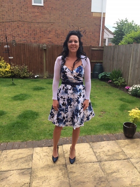 Sandra Millett, with long dark hair, stands on a patio in a garden, wearing a white and blue floral dress, a light shawl, dark heels, and a small fascinator. A wooden fence, lawn, and lush plants frame the scene.