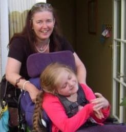Sandra Robertson stands smiling behind a girl in a wheelchair. The girl, with long blonde hair and a bright pink top, looks content. They are inside near a doorway, with keys hanging on the wall in the background.