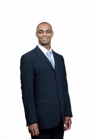 Stephen Ferguson, wearing glasses, a dark suit, a white shirt, and a light blue tie, stands against a plain white background, smiling at the camera.