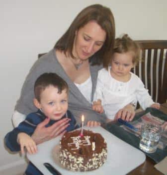 Suzanne Kaufman style shines through as a woman sits at a table with a young boy and toddler girl on her lap, smiling warmly as the boy prepares to blow out a candle on his birthday cake, celebrating together.