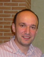 Blagojche Arsovski, a man with short hair and a receding hairline, smiles at the camera in a light pink striped shirt. A brick wall and a beige wall are visible in the background.