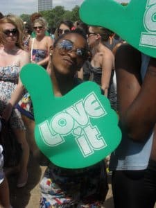 Natasha Ferguson, wearing sunglasses, smiles in a crowd whilst holding a large green foam hand that says LOVE it. Other people in summer clothes mingle in the background.