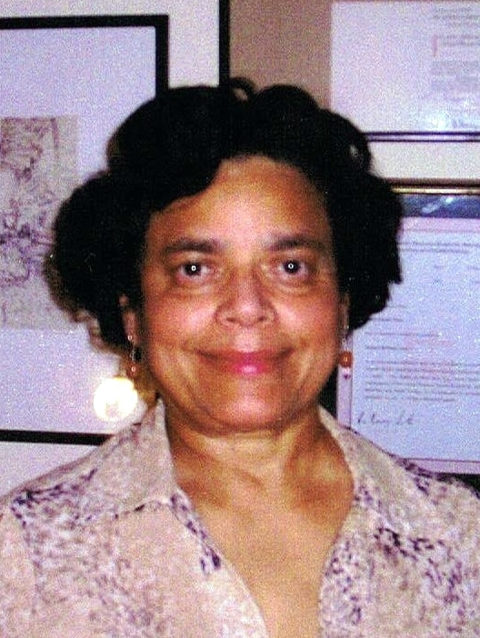 Premi Bonomally, a woman with short curly hair wearing a patterned blouse, smiles at the camera. She stands indoors in front of a wall decorated with framed pictures and documents.
