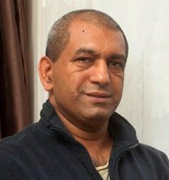 Shaf Sabir, a middle-aged man with short, greying hair and medium skin tone, is wearing a dark zip-up jumper while standing indoors in front of a light-coloured wall and a brown curtain.