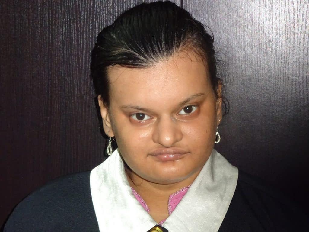 Satavisa Lahiri, with dark hair pulled back and silver earrings, is wearing a black and white outfit whilst standing in front of a dark-coloured wall, looking directly at the camera.