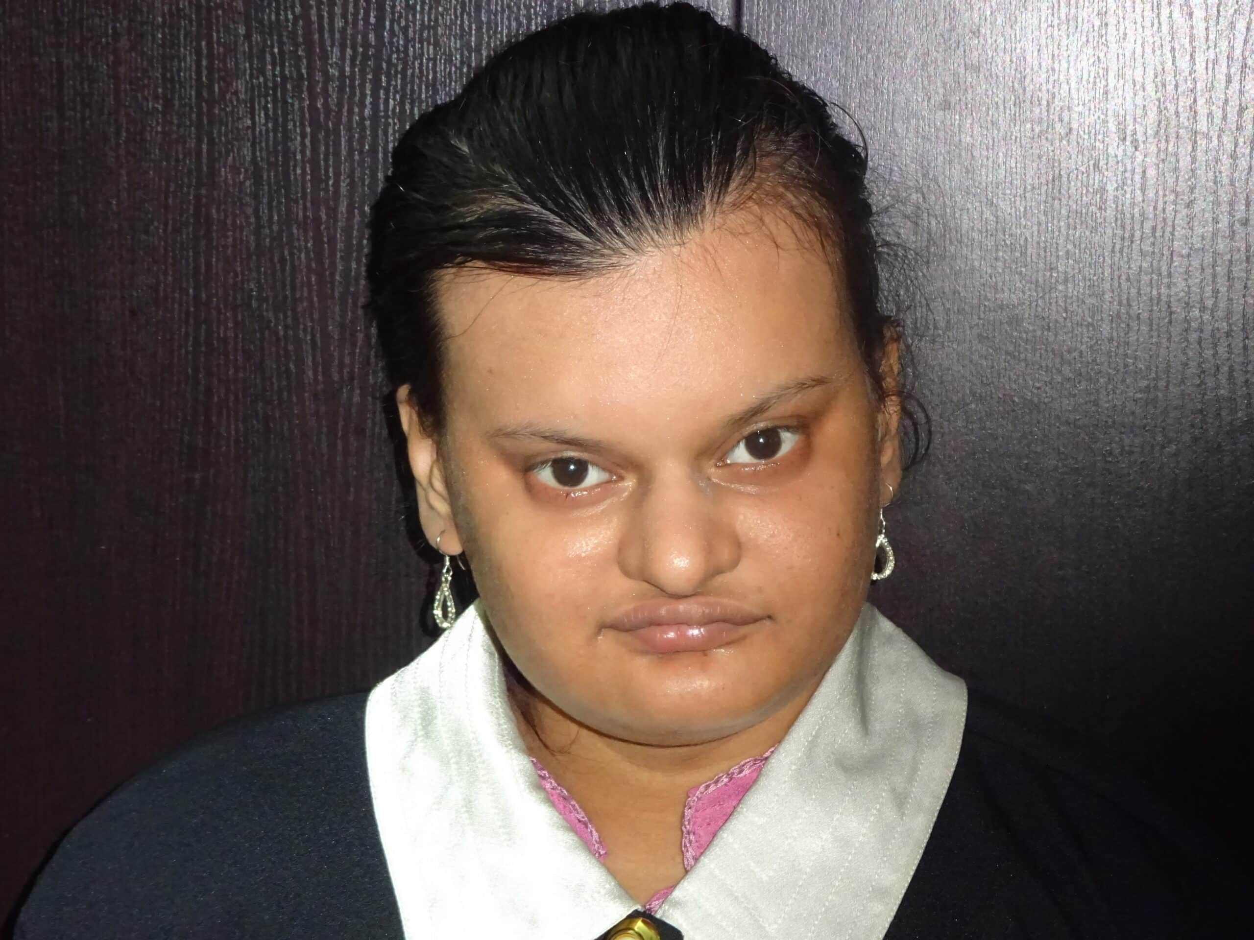Satavisa Lahiri, with dark hair pulled back and silver earrings, is wearing a black and white outfit whilst standing in front of a dark-coloured wall, looking directly at the camera.