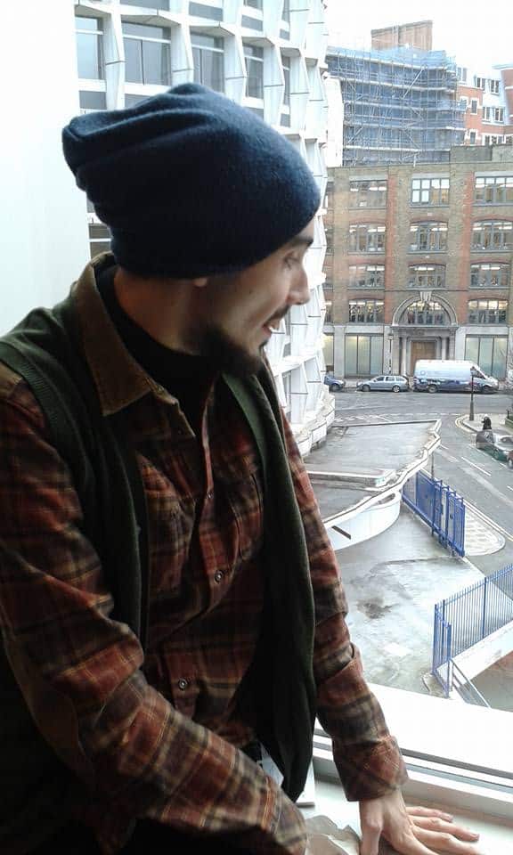 Roni Penny, wearing a blue beanie, checked shirt, and waistcoat, looks out of a window. Urban buildings, a curved drive, and blue gates are visible outside. Roni appears to be smiling or speaking.