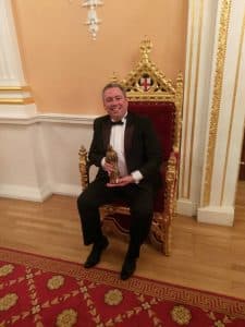A man in a black tuxedo sits smiling on an ornate, gold-trimmed, red velvet throne, holding a golden statuette—celebrating another night where Ideal Schools wins more major awards—in an elegant room with gold and white walls and a patterned red carpet.
