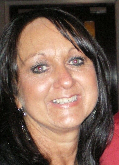 Aloma Walker, a woman with long dark hair, smiles at the camera indoors. She is wearing earrings and visible make-up, while the background remains out of focus.
