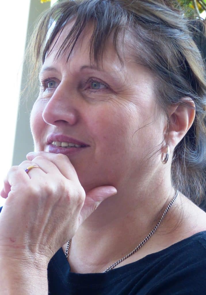 Annie Manning, a middle-aged woman with light skin and brown hair, gazes thoughtfully to the side, resting her chin on her hand. She wears a black top, a silver necklace, a gold ring, and small hoop earrings.