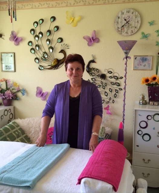 A woman with short brown hair, wearing a purple jumper, stands behind a massage table with folded blue and pink towels in a brightly decorated room with peacock and butterfly wall art, embodying Totka Lefedjiev - The road from science to alternative medicine.