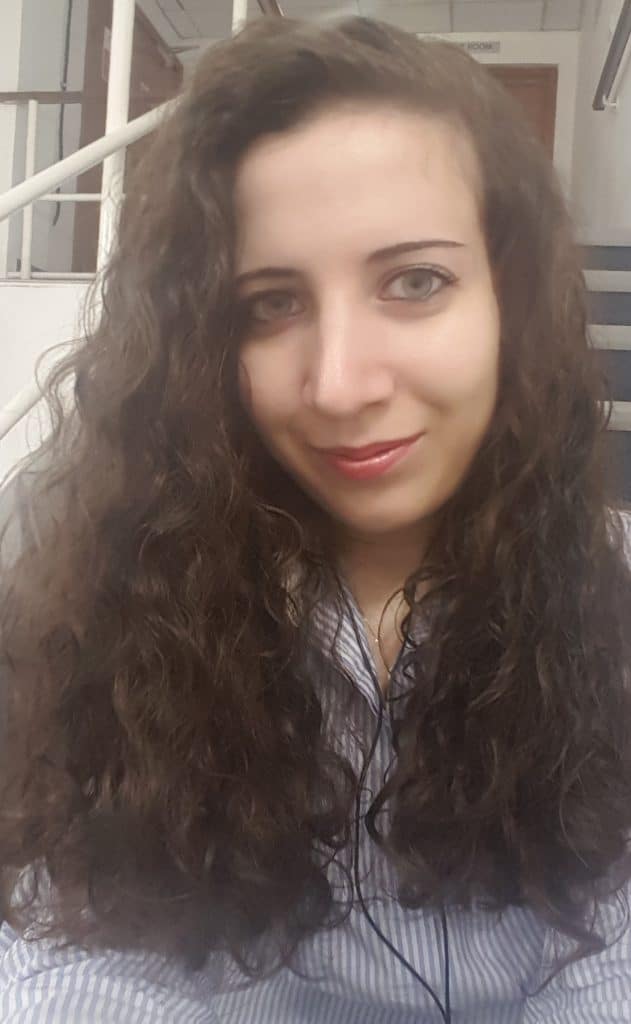 A woman with long, curly brown hair and a striped shirt smiles softly at the camera. Indoors, she sits near a staircase with a white handrail behind her. This could be Alessandra Lanzalaco captured in a quiet, candid moment.