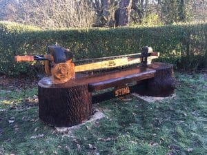 A wooden bench carved to resemble a large saw, with two axe heads as armrests and “STIHL” branding on the blade and bench front, designed by Steve Greenwell, situated on grass with trees and hedges in the background.