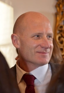 A middle-aged man with a bald head, wearing a white shirt, red tie, and dark suit jacket, smiles whilst looking to the side; ornate gold décor is visible in the background—this is Michael Panrucker.