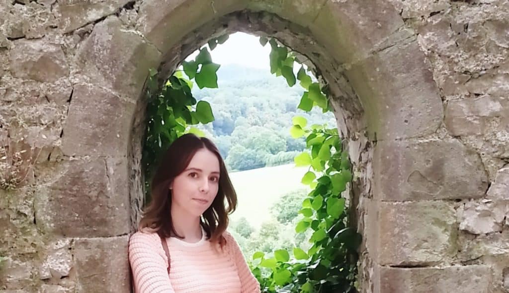 Cheryl Lewis, a woman with brown hair wearing a light pink jumper, stands in front of a stone archway covered in green ivy, with lush countryside visible through the opening.