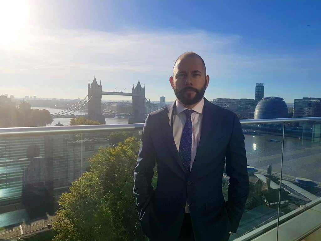 Chris Middleton, dressed in a suit, stands on a balcony overlooking the River Thames, with Tower Bridge and city buildings in the background under a clear blue sky.