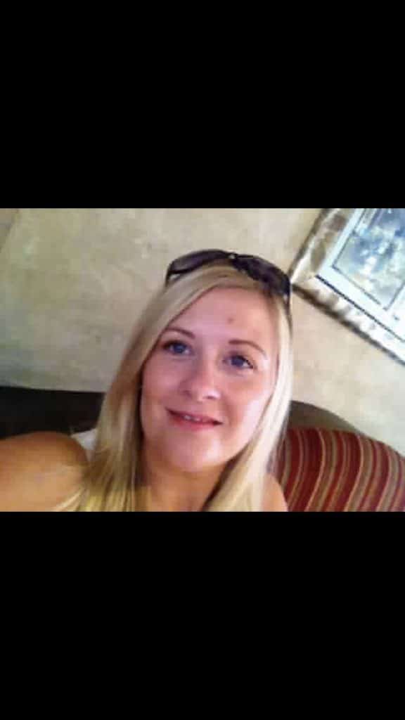 Jayne Tooke, with long blonde hair, sits on a striped sofa smiling at the camera. She wears sunglasses on her head and is indoors, with a beige wall and a framed picture in the background.