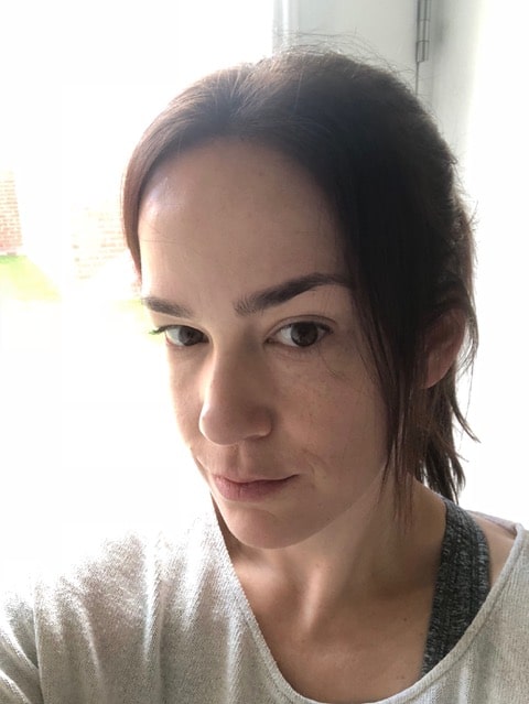 Lorena Diaz, a woman with fair skin and dark brown hair tied back, looks at the camera with a neutral expression. Indoors and wearing a light-coloured top, she is softly illuminated by natural light from behind.