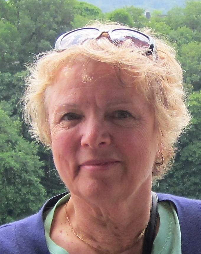 Eline Staplehurst, a woman with short, curly blonde hair, smiles softly at the camera. She wears sunglasses on her head and a dark top while green trees fill the blurred background.