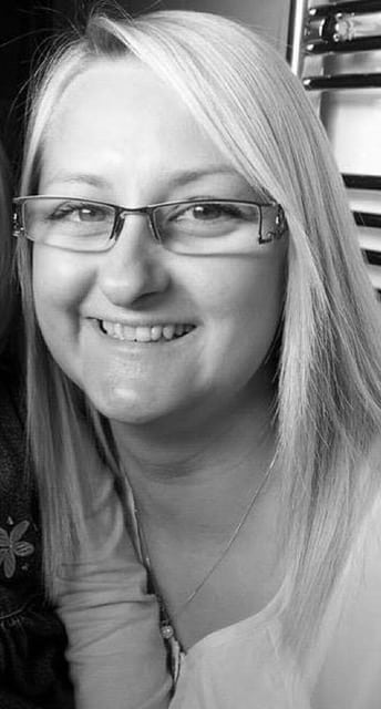 Amanda Frankland, a woman with straight, light-coloured hair and glasses, is smiling at the camera. She is wearing a light-coloured top and a necklace. The black-and-white photo captures her warm expression.