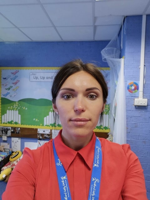 Anna Shutyak, a woman with brown hair, wearing a red shirt and blue lanyard, stands indoors in front of a classroom display board featuring a nature scene with birds. The blue wall behind her is decorated with a colourful ball.