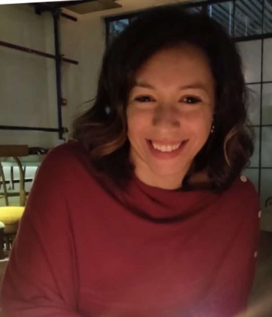 Dalia Abdel-Salam, a woman with wavy brown hair wearing a red top, smiles warmly whilst sitting indoors at a dimly lit restaurant or café.