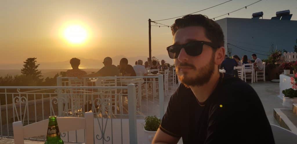 Jamie Munro, wearing sunglasses, sits at an outdoor restaurant during sunset, with other people dining in the background and a scenic view of the sun over the horizon.