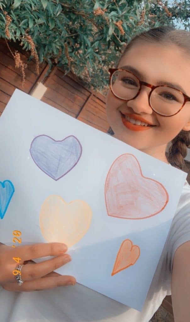 A smiling person with glasses, possibly Mia Blakemore, holds up a white sheet of paper featuring colourful, hand-drawn hearts. There is greenery and a wooden fence in the background, with a date stamp reading 20/4.